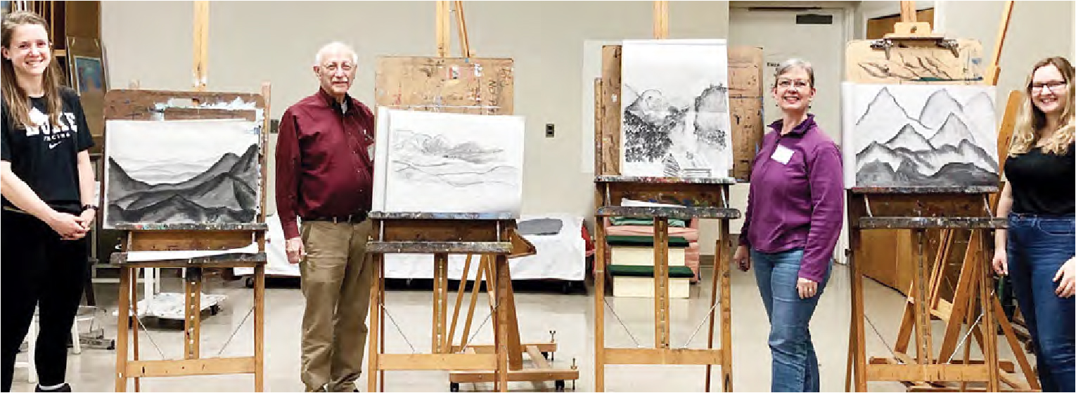 Four students stand next to their pencil drawings of a mountain scene smiling at the camera