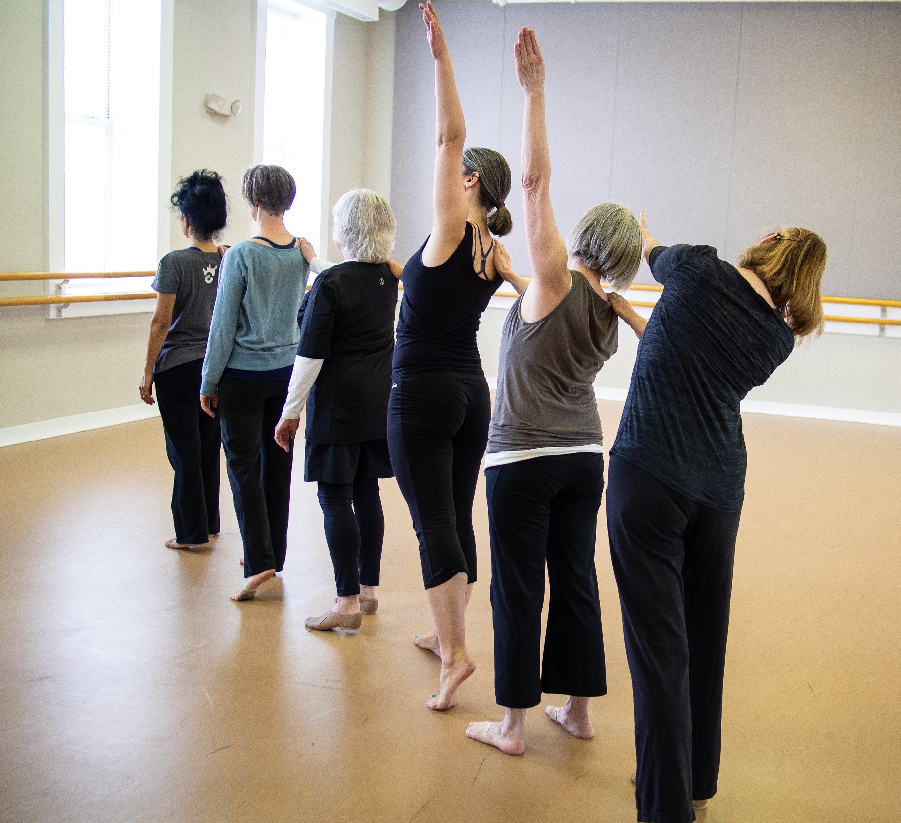 6 women in a diagonal line facing away from camera rehearse in a dance studio